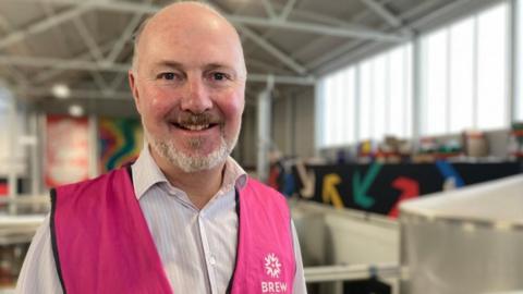Peter Wells, standing inside a brewery, wearing a pink shirt, with stripes and a pink high-vis jacket. He is smiling at the camera, with signs and equipment behind him. 