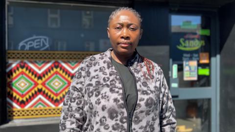Shows a woman with her hair in plaits and a cardigan standing in front of a restaurant 