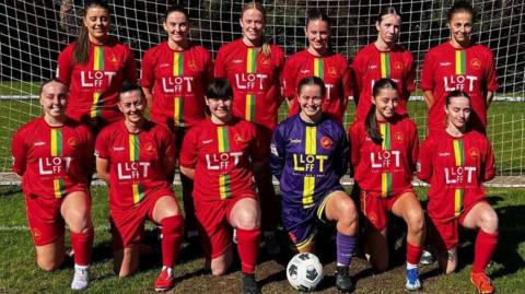 A group of young women from the CPD Merched Y Felinheli team, all dressed in red football kits - bar the goalkeeper, who is in blue - formally arranged in front of a goal.