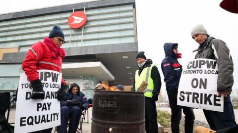 Striking union workers stay warm around a fire burning in a barrel, carrying signs that read "CUPW Toronto Local On Strike"