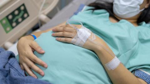 A pregnant woman waits to give birth lying in a hospital bed, wearing a blue gown and mask, with a tube attached to the back of her hand