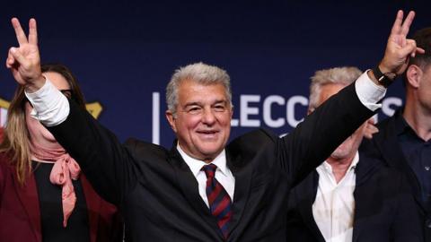 Joan Laporta raises his hands in celebration after winning the Barcelona presidency election. He has grey hair and is wearing a dark suit, white shirt and a tie in Barcelona's club colours - red and blue.