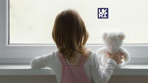 Young girl looking out of the window holding a teddy bear