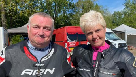 Dave and Alison looking into the camera and smiling. They're both wearing black motorbike leathers. Dave has short grey hair with sideburns and is wearing a blue undershirt. Alison has short blonde hair and is wearing a pink undershirt. They're stood in front of their red van, which has 'Terrier Racing' printed on the rear window. 