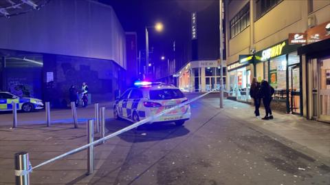 A police cordon along a dark street. Two police cars are in the image - one with flashing blue lights.