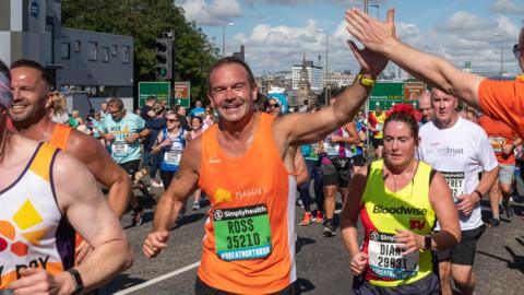 Ross running a half marathon, surrounded by a group of people also running. He has short brown hair and is wearing an orange vest with a race number pinned on. He is high-fiving a person from the crowd. 
