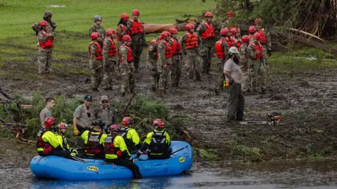 Rescue teams wearing yellow and orange fluro clothes on a muddy river bank and sitting in a blue dingy on a river.