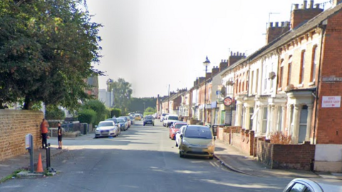 A residential street, with cars parked by the side of the road and a row of terraced houses to to the right.