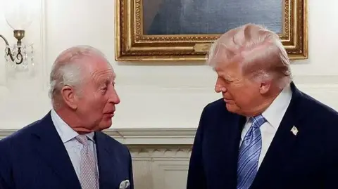 King Charles, left, speaks to Donald Trump, with both men turned toward each other in front of a portrait of George Washington.
