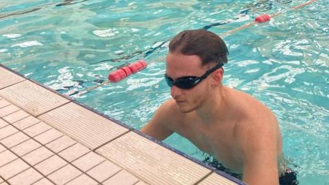 Robert Constantin with short dark hair wearing swimming goggles as he gets into an indoor swimming pool with a light brown surround and blue water. A lane rope is visible alongside.