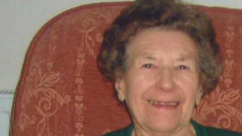 Una Crown smiles for the camera. She is sitting on a pink-brown armchair and is wearing a green top.