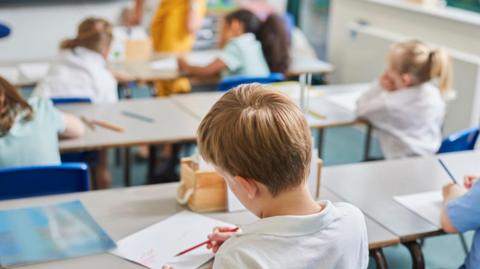 A group of children in a classroom sitting on blue chairs with their backs to the camera. They are wearing either white shirts or green dresses and have paper and pencils on their desks. 