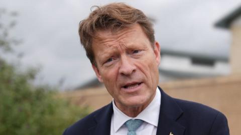 A head and shoulders shot of a man wearing a blue suit jacket, shirt and tie. He has brown wavy air and is standing outside a building, which is blurred out in the shot.