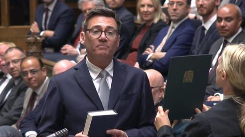 Andy Burnham holding a Bible being sworn-in as an MP in the Houses of Commons. He is wearing a dark blue suit and white shirt.