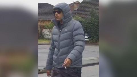 A CCTV image of a man wearing sunglasses and a hooded coat while smoking a cigarette