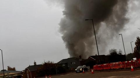 Thick grey smoke travelling from a building that is on fire, there are red plastic traffic cones and barriers on the side of a junction.