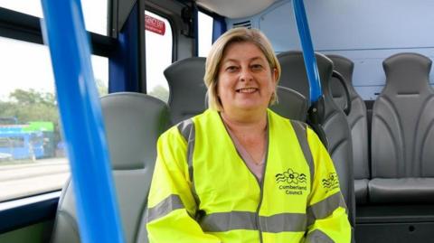 Councillor Denise Rollo smiling into the camera. She has a blonde bob and is wearing a yellow hi-vis jacket with Cumberland Council's logo on the breast. She is sitting on a bus with grey seats.