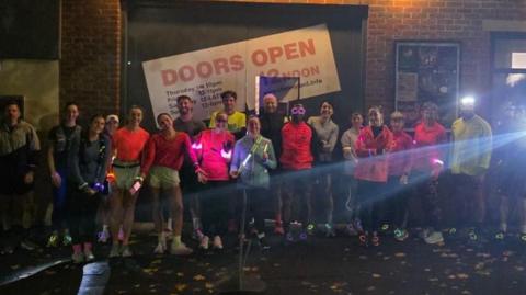 Night-time shot of several members of the run club. They're wearing multi-coloured running gear and standing in a line for the photo.