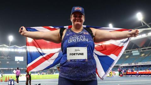 Sabrina Fortune smiling and holding the Union Jack flag behind her back with her arms outstretched, celebrating her victory.