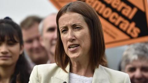 Jane Dodds in a cream suit. There are people stood behind her holding Liberal Democrats signs suggesting the photo was taken at a campaign rally.