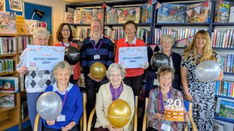 Nine members of the community library celebrating the 20 year anniversary with balloons, posters and a cake.