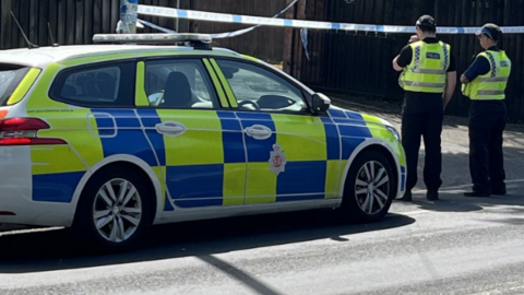 A police car and two police officers are stood outside crime tape which is in front of a large gate, in a residential area. 