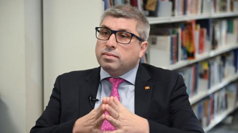 Mark Arnull in a navy blazer, blue shirt and pink tie sits in front of a book shelf looking off to the left of camera.