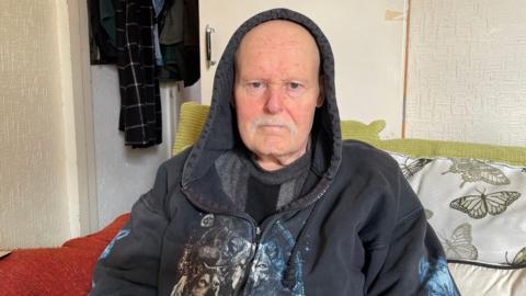 A man sits on a sofa looking directly at the camera with a serious expression. He is wearing a dark hoodie with an animal print design and has a grey moustache. Behind him are colourful cushions, a butterfly-patterned throw and coats hanging on the back of a door.