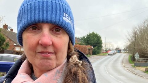 A woman with a blue hat, pink scarf and a blue coat standing by the side of a long main road