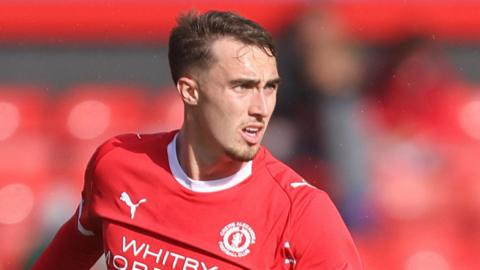 Lewis Billington in action for Crewe. He is wearing a red home shirt and is looking to his left preparing to pass the ball