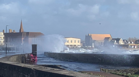 Waves can be seen overtopping the barrier at Troon. The sky is grey and sea mist is being blown high by the wind.