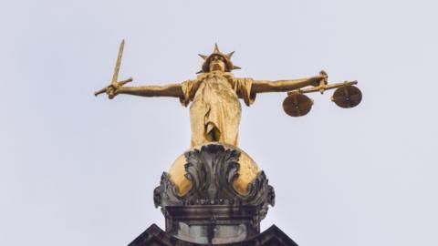 A gold statue on top of the Old Bailey from below. It is of a woman in a dress with a spikey hat and outstretched arms. In one hand she is holding some scales and the other a sword. The sky above is grey.