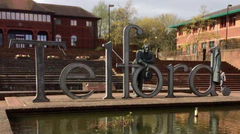 A metal sign spelling out the letters to T-E-L-F-O-R-D stands on a paved area next to a small pond. Behind it are stone steps leading to brick buildings. Over the letter "o" of the sign, leans a sculpture of a man.