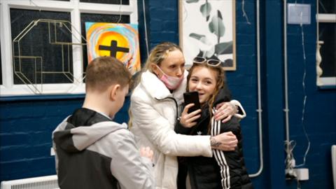 A woman in a white coat and medical mask hugs a teenager in a black tracksuit top with white lines on the sleeve. She has sunglasses on her head and is holding a mobile phone. Next to them is a teenage boy with a grey and black coat with his back to the camera. They are in a hall with blue walls