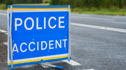 A blue and white sign, positioned on a country road, reads "Police Accident".