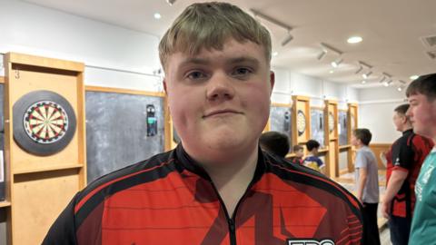 Young person in red-shirt with darts board behind.