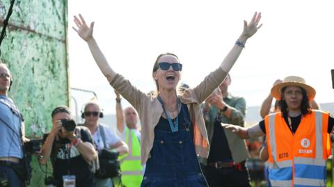 Emily Eavis opening the gates to the festival. She is talking with her hands held up in the air, as people film and take pictures behind her. 