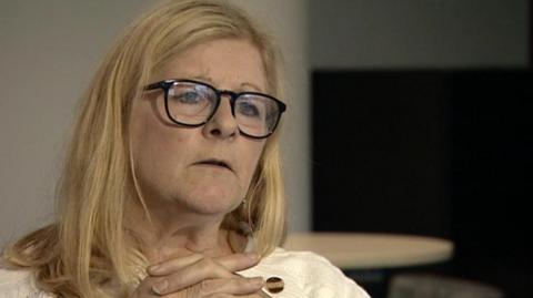 A woman with blonde hair wearing glasses with her hand crossed at collarbone level. She has black-rimmed glasses and sits in a room with white and dark walls and a round, wooden table behind her.