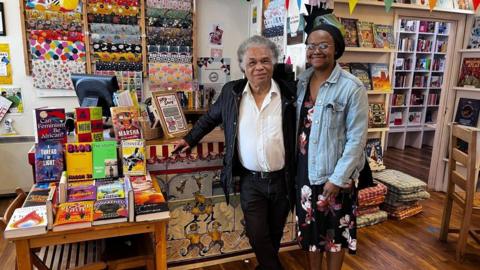 A man and a woman are stood in the middle, in front of a cashier desk in a bookshop. The man is on the left and has grey hair. He is wearing a white shirt, unbuttoned at the collar, and a black jacket, with black trousers and a belt. He is leaning against the desk with his right arm. The woman on the right is wearing a black hair bonnet, and is wearing a black dress with red and white floral decorations and a light blue denim jacket. To their left, there is a table displaying a colourful selection of books. There is a computer and more books on the desk, and behind the desk there is a display of wrapping paper hung on the white wall. There are more book shelves behind the two people, on their right.