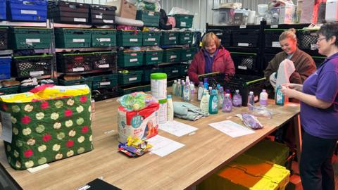 A photo showing three women stood around a table packing items into bags. In the background are trays of items stacked up