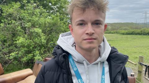 A young man with blond hair stands outside, looking into the camera lens. He is wearing a grey hoodie and navy coat, as well as a light blue lanyard which says "We're Kirklees" on it. Behind him is a pile of brown sofas, and a luscious green landscape in the background.