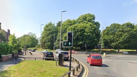 A road junction with traffic lights and a grass verge to the left, there are several cars including a small red one and two parked cars at the side of the road. 