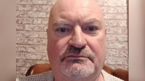 A selfie of a man with a grey beard and bald head against a white-washed brick wall.