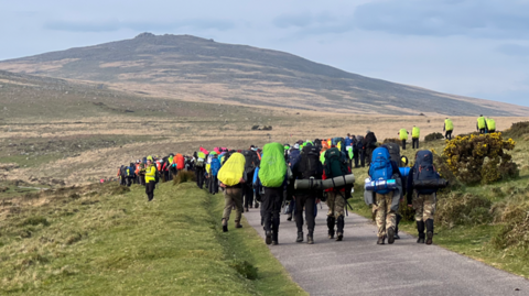 A large group of walkers set off with backpacks and camping gear. They are walking along a path on the moors on a grey day. 