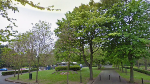 Tall trees line two concrete paths in a large park. There are bollards stopping traffic on one path and a car park can be seen on the left of the photo.