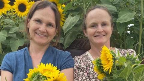 Two women in their late forties who share a family likeness sit together holding sunflowers and smiling. 