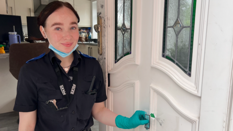 Woman in a police uniform standing in front of a door holding a clear sheet with a green arrow on it. 