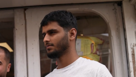 Mr Ali is a young man with a short black beard. He is wearing a white t-shirt and is standing in front of a shop