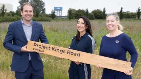 Three people stand in a grassy field holding a long wooden sign which reads: "Project Kings Road". The dark haired man on the left of the picture is wearing a navy blue suit with a striped shirt. A woman standing in the middle has dark hair and is wearing a navy co-ord set with white stripes down the side. A woman with blonde hair stands on the right of the image and she is wearing a navy dress with a crown broach.