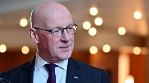 John Swinney, a bald man with glasses, wears a dark suit, white shirt and purple tie. He is looking to his left in a medium-close up shot in the Scottish Parliament, with twinkling lights in a blurred background 